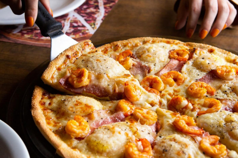 Foro da mão de uma mulher pegando uma fatia de uma pizza de camarão, da Pizza Bis, em Balneário Camboriú. 