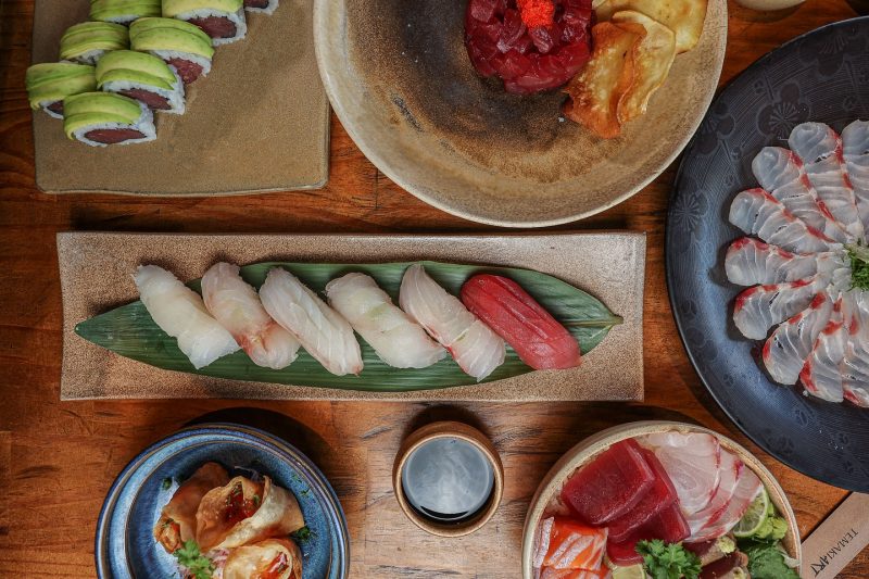 Foto tirada de cima de vários pratos orientais dispostos em louças de diferentes formatos geométricos e cores sobre uma mesa de madeira. Os alimentos são do restaurante Temaki Art, em Balneário Camboriú. 