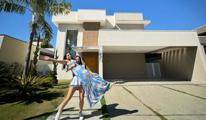 Mulher em pé carrega outra nos braços em frente a uma casa de luxo bege, sob céu azul
