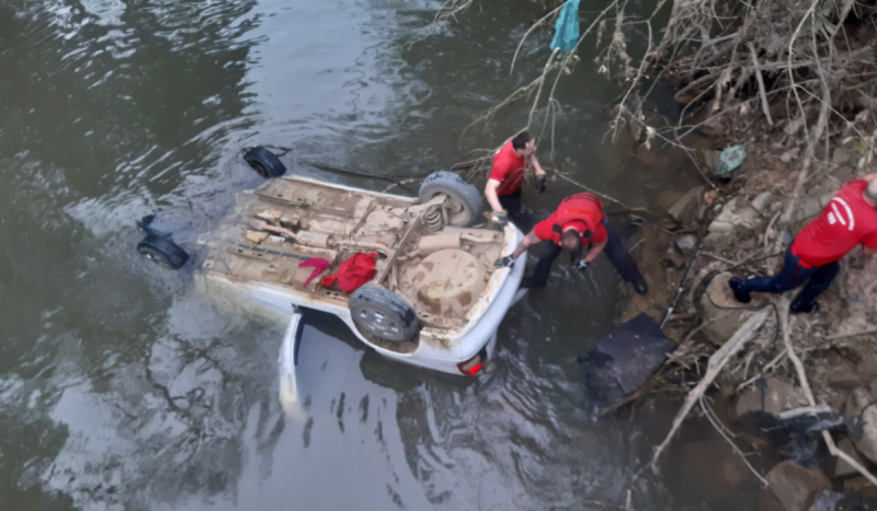 Motorista morreu após carro branco cair de ponte em SC. Foto mostra carro branco com as rodas para cima dentro de rio parcialmente submerso