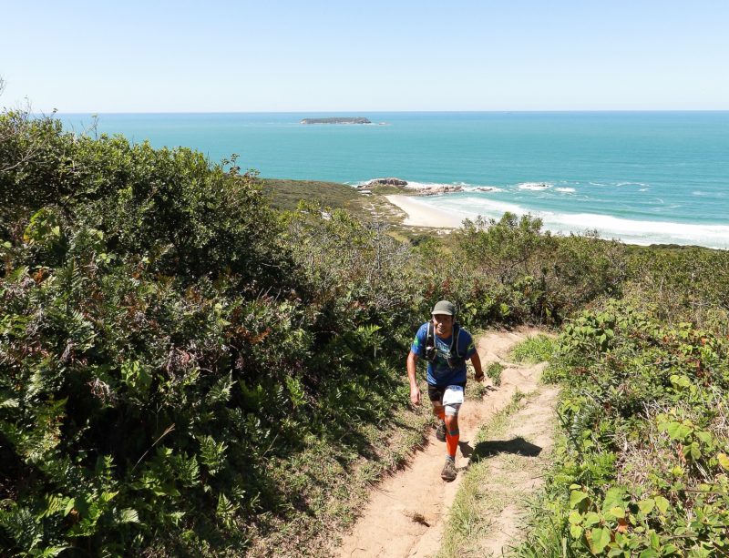 Foto de um homem corredor subindo uma trilha de terra em cima de um morro em Florianópolis. Ao redor há alguns arbustos e vegetação rasteira e ao fundo, o mar e o céu azul. O dia está ensolarado. 