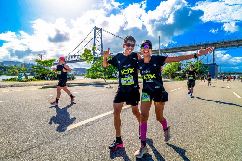 Foto de um homem e uma mulher corredores de rua. Eles estão lado a lado e com uma de suas mãos esticadas. Eles sorriem para a foto. Ao lado e ao fundo, aparecem outras pessoas participando da corrida de rua. Ao fundo, a Ponte Hercílio Luz e o céu azul. O dia está ensolarado. 