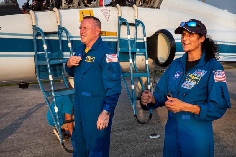 Astronautas Suni Williams e Butch Wilmore com roupas espaciais por tras de nave