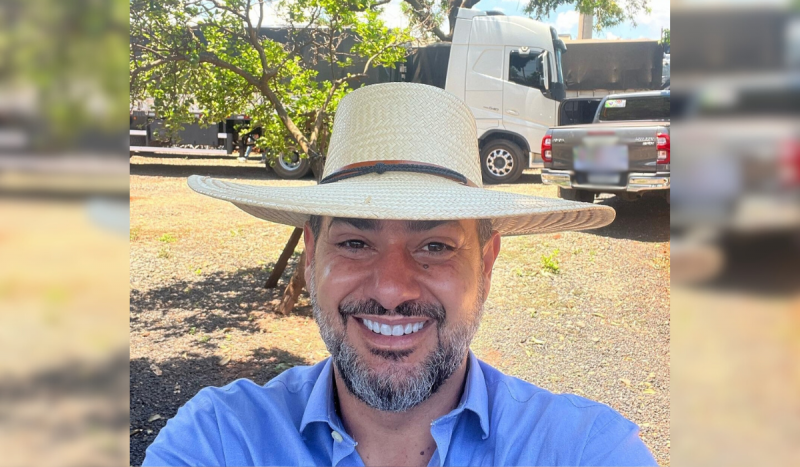 Homem branco usando barba e bigode, sorri enquanto usa chapéu de palha em meio à propriedade rural. Atrás dele aparece um caminhão e uma caminhonete cinza