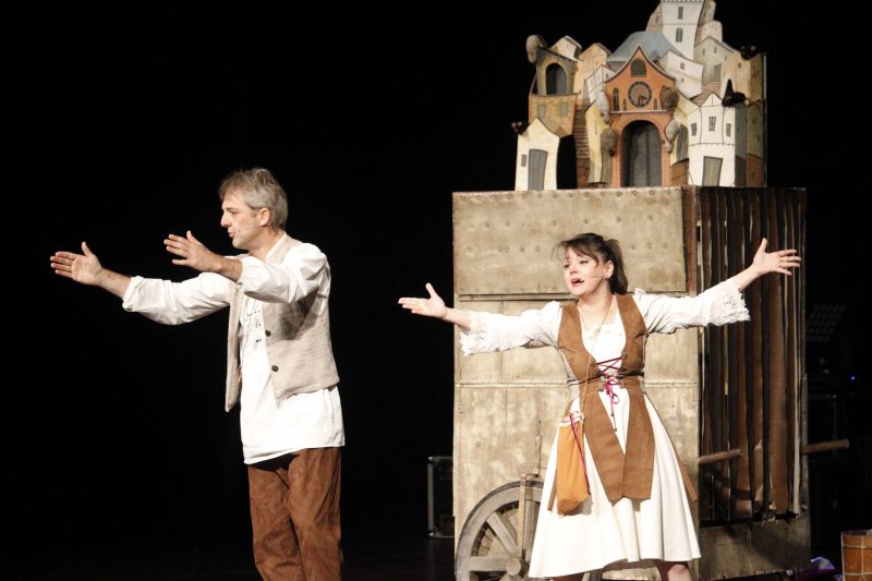 Homem e mulher de meia idade, vestidos com roupas nas cores branca, bege e marrom com braços abertos. Eles estão em uma apresentação teatral. Ao fundo, uma estrutura de madeira sobre a qual há pequenas casas, como cenário. 