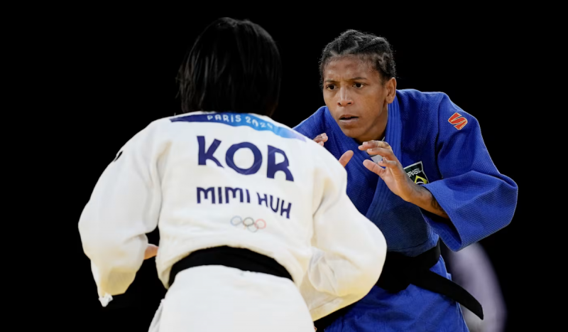 Judocas lutando nas Olimpíadas de Paris 2024, de costas para a foto, uma mulher de cabelos curtos e escuros veste quimono branco e, de frente para a foto, atleta negra veste quimono azul