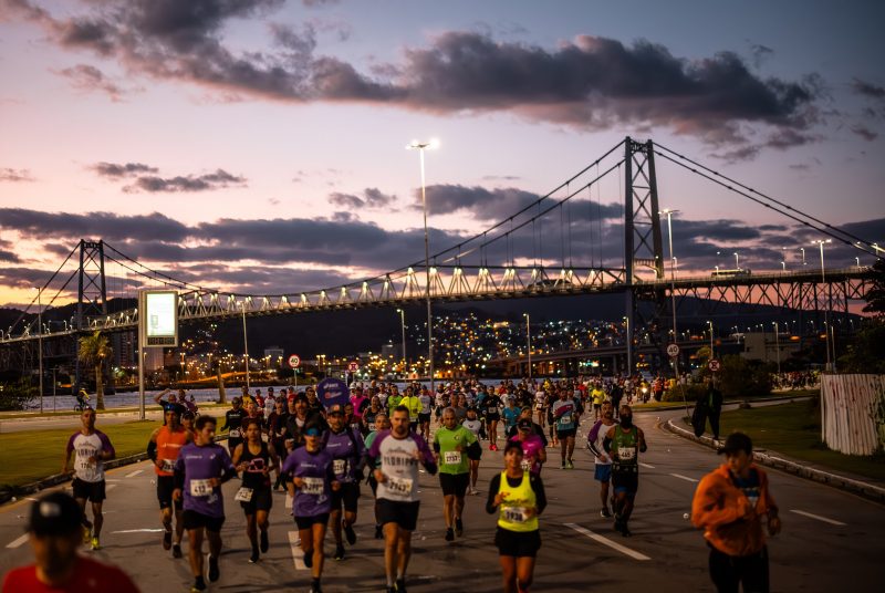 Foto de diversas pessoas correndo em uma via de Florianópolis. Ao fundo, a Ponte Hercílio Luz e atrás dela, um morro com luzes acesas de casas. A foto foi tirada ao anoitecer. 