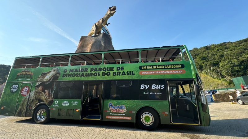 Foto de um ônibus turístico de dois andares. Ele é plotado com uma estampa verde e uma foto de um dinossauro com a boca aberta. Ao lado, está escrito: O maior parque de dinossauros do Brasil. Em cima do ônibus tem uma escultura de um dinossauro em cima de uma rocha. 