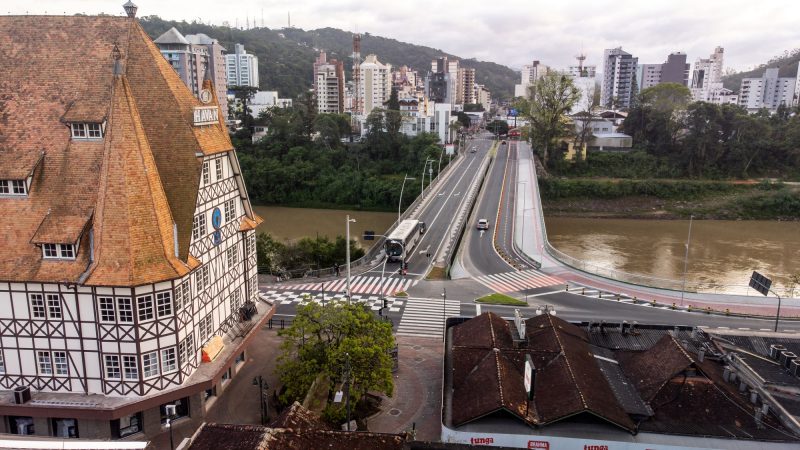 Foto aérea do Centro do Centro de Blumenau. No lado esquerdo da foto, o castelinho em estilo germânico onde fica a loja Havan. No meio da foto, uma ponte de asfalto que passa por cima do Rio Itajaí Açu. Ao fundo, vários prédios altos. 