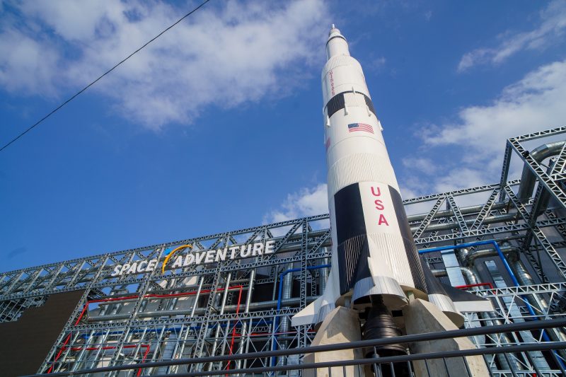 Fachada do Parque da Nasa. A foto foi tirada de baixo para cima e até a metade da imagem aparece a grande estrutura metálica com o nome Space Adventure. À frente dela, uma grande réplica de foguete branco, com a bandeira dos Estados Unidos e a sigla USA. Acima, o céu azul. 