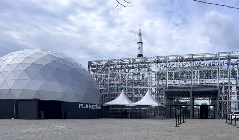 No lado esquerdo da foto, o planetário. Ele é uma estrutura grande e de cobertura arredondada e prata. No lado direito, aparece as costas da bilheteria do parque, uma grande estrutura metálica onde está escrito Space Adventure. Atrás dela aparece um pedaço da réplica de foguete que fica em frente à fachada do parque. 