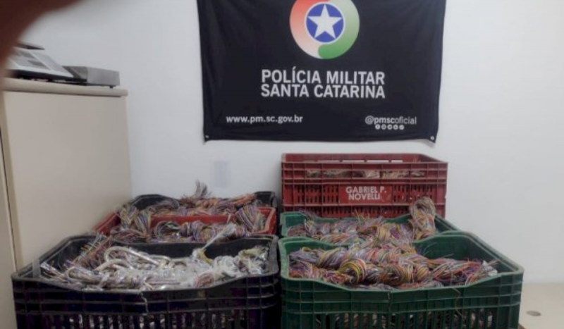 Caixas coloridas de plástico com sacos de fios de cobre e atrás uma bandeira com a insígnia da Polícia Militar de Santa Catarina