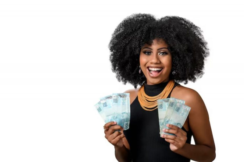 mulher negra com cabelo encaracolado, sorrindo, com uma blusa preta e um colar de cor laranja, segurando nas duas mãos leques de notas de cem reais 