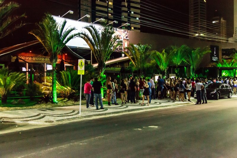 Foto noturna de uma balada na Avenida Atlântica. Várias pessoas estão na calçada em frente ao estabelecimento. Em frente à fachada, que não aparece direito, há seis coqueiros iluminados com holofotes verdes.