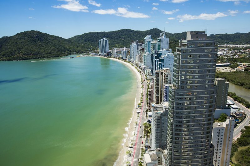 Vista área da Praia Central de Balneário Camboriú. Do lado esquerdo ao meio da foto, o mar esverdeado e a areia da praia. Do lado direito, a Avenida Atlântica e os altos prédios. Ao fundo, morros cobertos por vegetação.