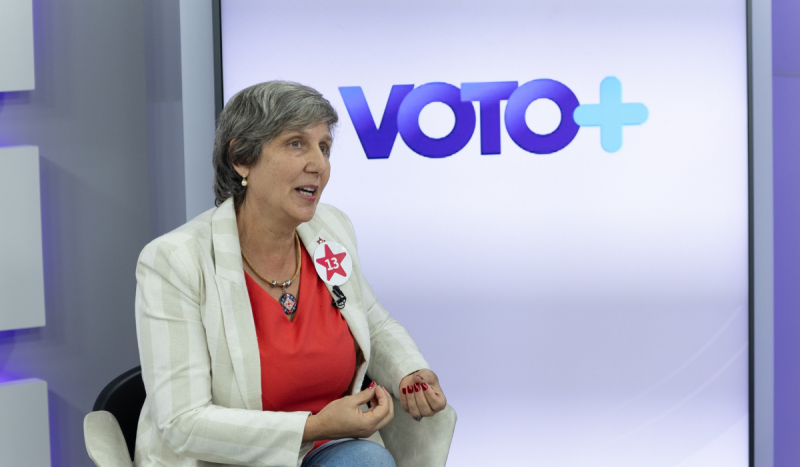 Tânia Slongo, candidata do PT a prefeita de Palhoça - mulher branca, de cabelos curtos e grisalhos, usando blusa vermelha com decote V, e blazer bege. Ela está sentada gesticulando com as mãos sobre as pernas em frente a um telão com o escrito 'Voto+'