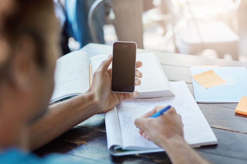 Pessoa com celular na mão realizando anotações em um caderno