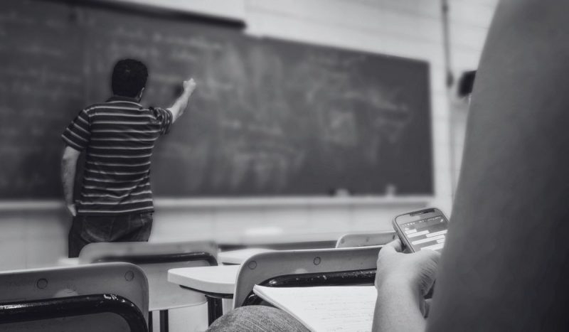 tradicional colégio de Florianópolis foi condenado após demitir professor que recebeu encontrou bilhetes com xingamentos homofóbicos na sua mesa durante a aula, foto mostra professor escrevendo no quadro enquanto aluno mexe no celular