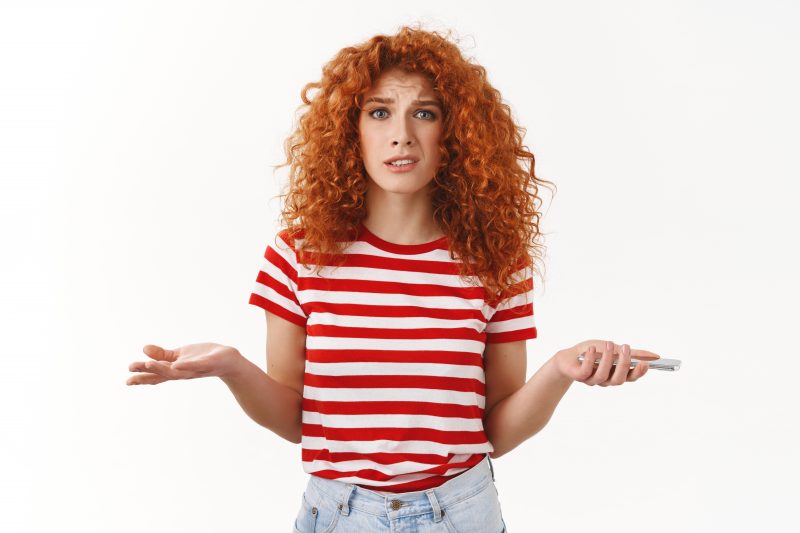 Mulher ruiva, com cabelos cacheados até a altura do ombro, usando uma camiseta com listras vermelhas e brancas e uma calça jeans, a foto é acima da cintura dela, ela está com um celular na mão esquerda e fazendo sinal de dúvida com as duas mãos, a expressão dela também é de dúvida com algo