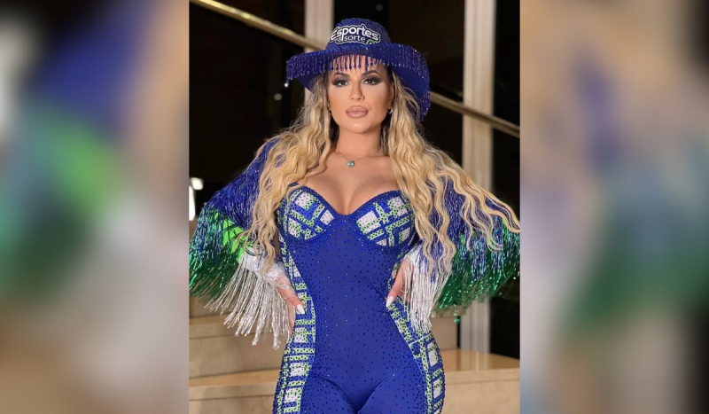 Deolane Bezerra é uma das pessoas que divulga apostas online, como Jogo do Tigrinho - mulher branca, em pé, usando roupa azul com franjas nos braços. Ela usa chapéu de boiadeira azul, também com franjas