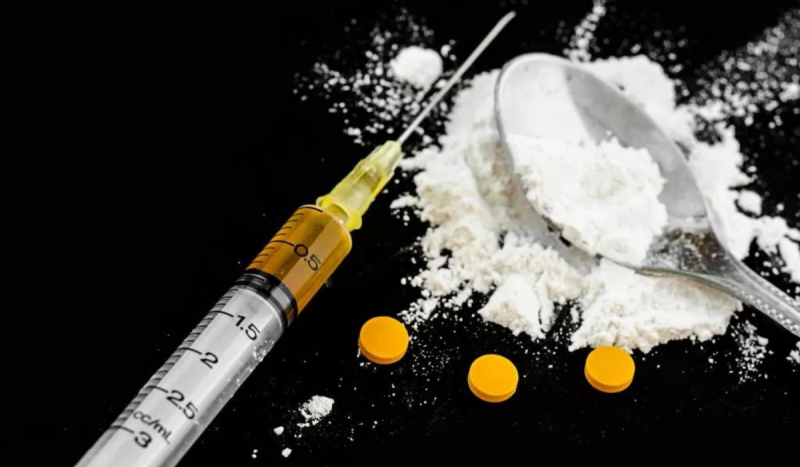 Droga zumbi - foto mostra seringa com ahulha com 0.5 ml de líquido amarelo. O item está disposto ao lado de uma colher com pó branco derramado em cima de uma superfície preta