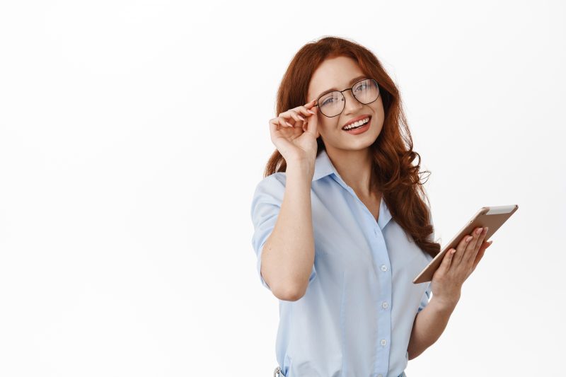 mulher ruiva com cabelo na altura dos ombros, camisa social de botão azul claro dobrada até o cotovelo, segurando na mão esquerda um tablete e com a mão direta segurando a lateral do óculos que está no rosto da mulher, ela está sorrindo e com o rosto levemente inclinado para a direita, indicando uma mulher extremamente inteligente