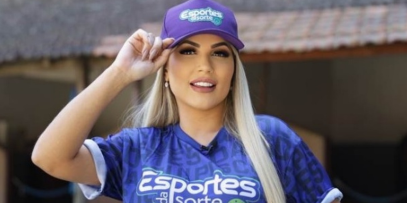 Mulher branca e loira de cabelos lisos compridos usando chapéu e camisa em tons de roxo e azul. Ela é Deolane Bezerra investigada em operação de jogos ilegais