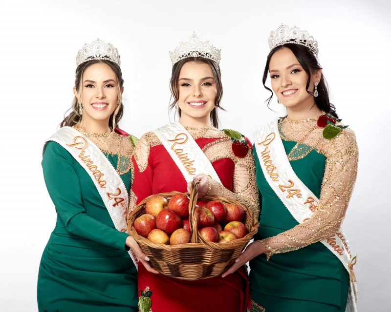 A foto mostra três mulheres jovens, de pele clara, cabelos cumpridos e presos. Elas estão uma ao lado da outra e seguram uma cesta de vime cheia de maçã. Cada uma usa uma tiara prateada na cabeça. As duas mulheres da ponta usam um vestido longo verde com renda dourada no braço esquerdo e um bordado de maçã na altura do ombro. Elas usam uma faixa onde está escrito: Princesa 24ª Festa Nacional da Maçã. A mulher do meio usa um vestido no mesmo estilo, mas na cor vermelha. Na faixa dela está escrito Rainha 24ª Festa Nacional da Maçã. 