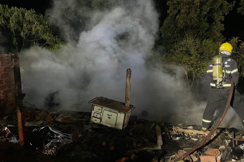 local onde era a casa consumida pelo incêndio, na imagem aparece um bombeiro de costas apagando o fogo com uma mangueira de água e ao lado um fogão a lenha que sobrou após as chamas