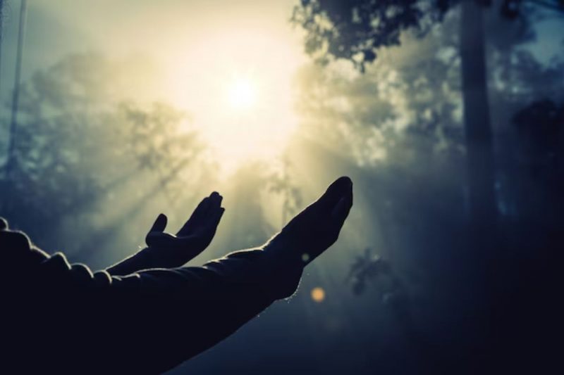 pessoa com as mãos estendidas para o céu em meio a floresta e com uma luz amarela ao fundo 