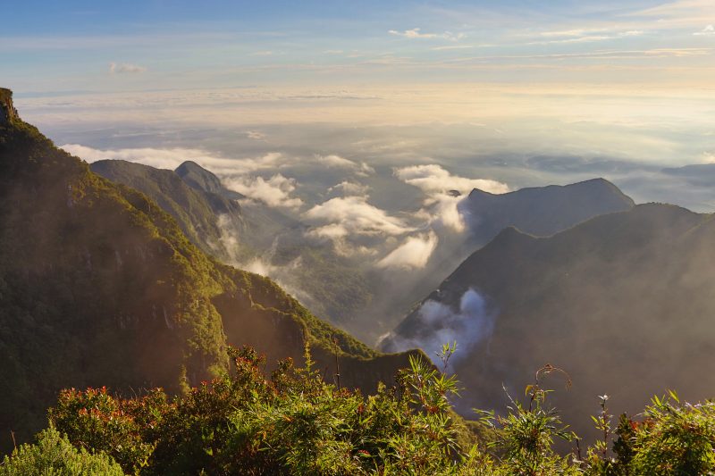 Foto panorâmica da serra com morros e nuvens por entre os cumes. O céu está claro e o dia ensolarado. 