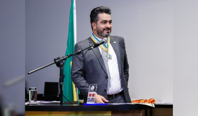 Homem branco usando camisa social branca e terno cinza, de topete e cabelos pretos, discursando em microfone