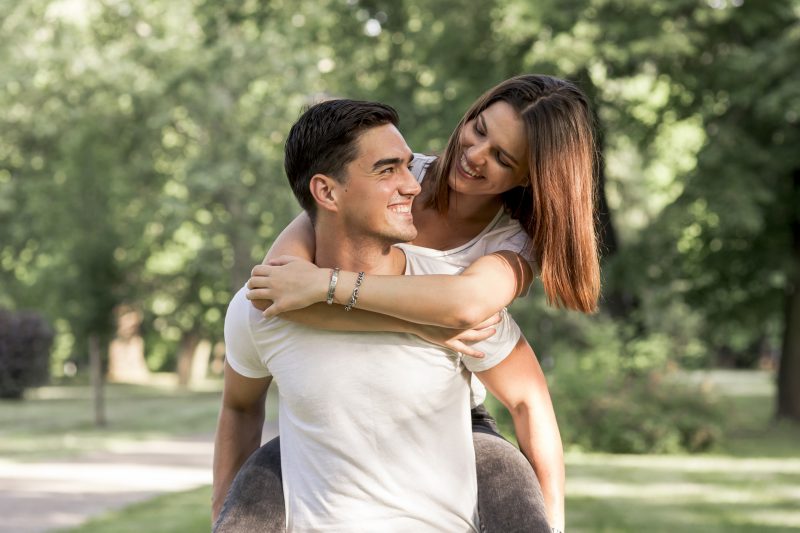 casal sorrindo; homem segurando a mulher nas costas e olhando para ela; ele está com uma camiseta branca e tem o cabelo preto e curto, a mulher está com uma calça preta desbotada, uma camiseta branca e tem cabelo curto e castanho; eles estão em um parque com muitas árvores 