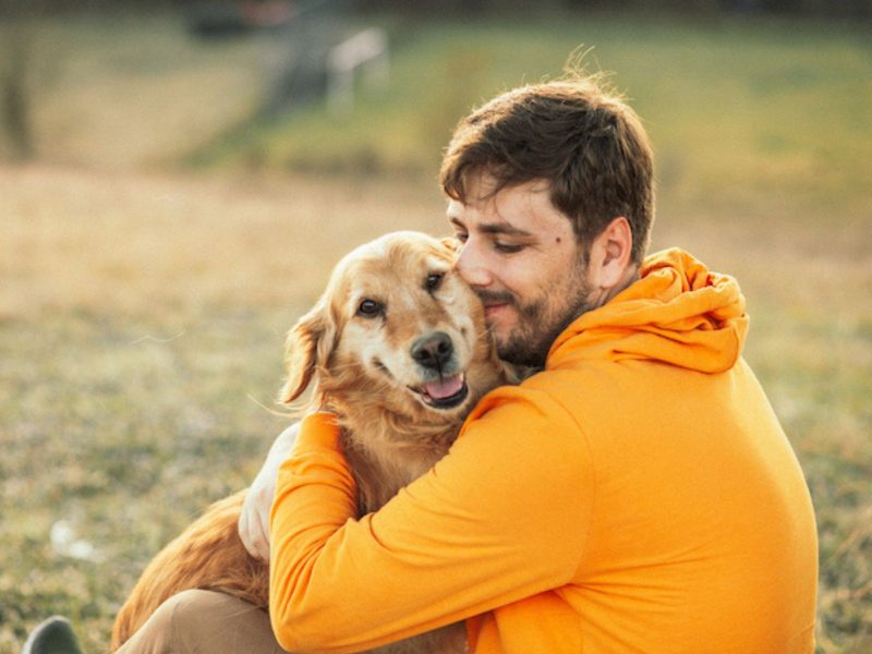 A imagem mostra um cachorro abraçado ao seu dono que está usando uma camiseta de mangas longas laranja.