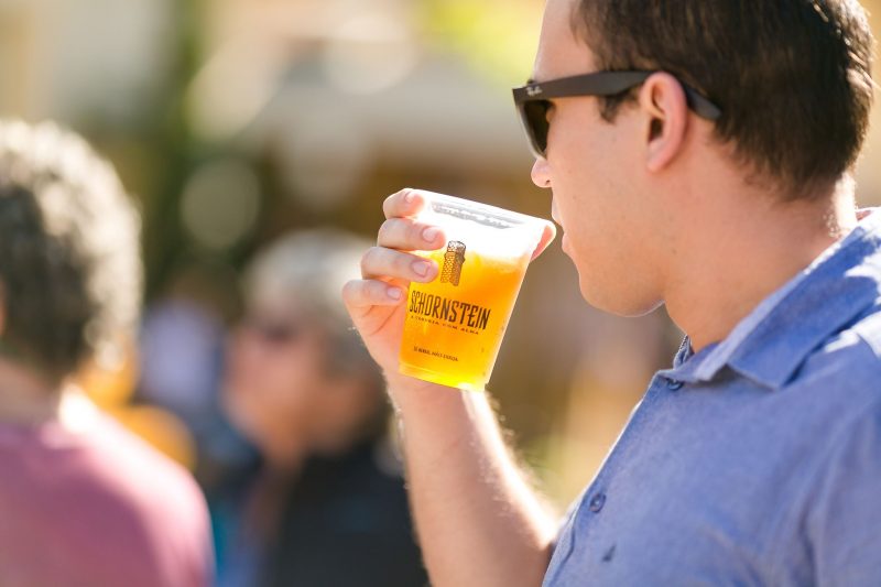 Foto de perfil de um homem jovem, de pele clara, segurando um copo plástico com chope e a logomarca da cervejaria Schornstein. Ao fundo, algumas pessoas desfocadas.