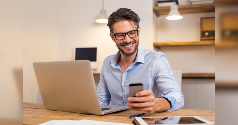 Homem sorrindo para celular na frente de computador