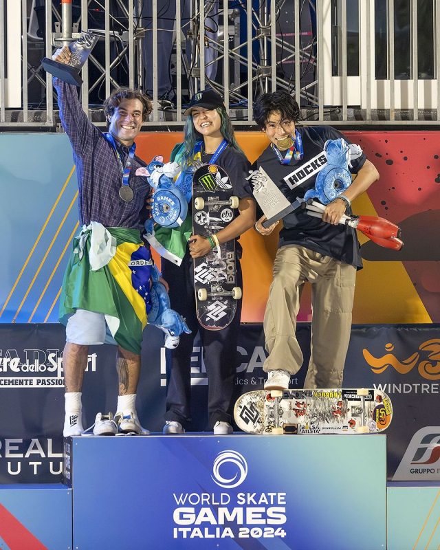 Pedro Barros, enrolado na bandeira do Brasil, ao lado de Raicca Ventura, com skate na mão, e augusto Akio