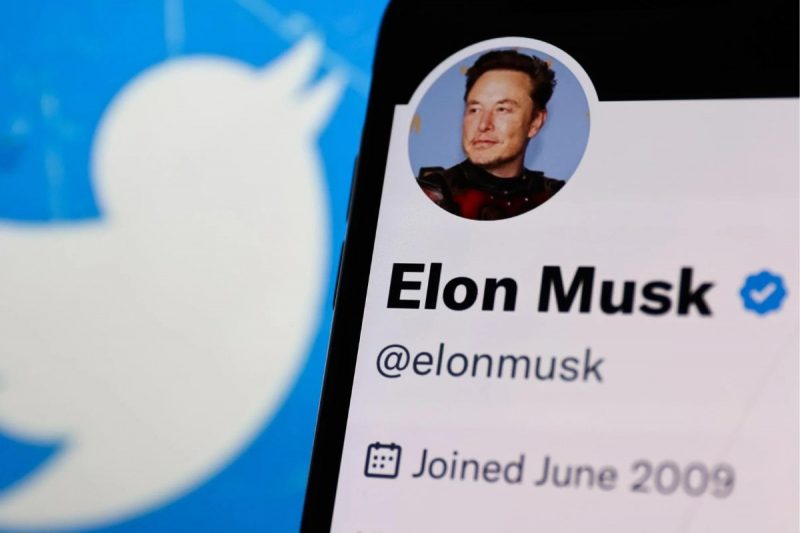 Imagem de um celular logado na conta de elon musk no twitter, com o antigo símbolo da rede social no fundo 