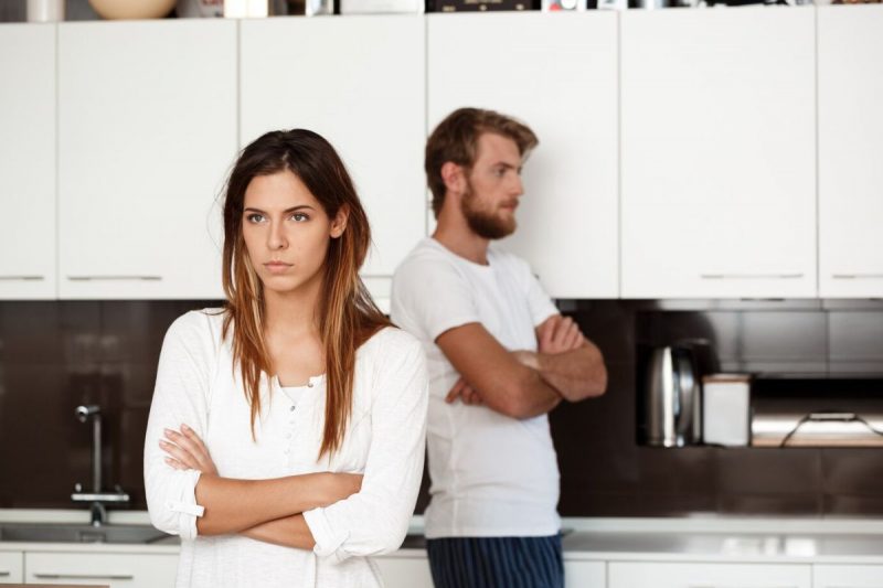 casal após uma discussão e suposta separação emburrados um com o outro; a mulher em primeiro plano de blusa branca, braços cruzados e a cara fechada e o homem ao fundo de braços cruzados e emburrado