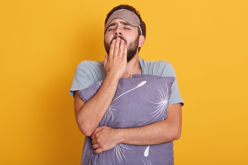 homem de camiseta cinza, segurando um travesseiro de fronha roxa com detalhes em branco e com um tapa olho na testa, ele está com a mão na boca bocejando signos preguiçosos