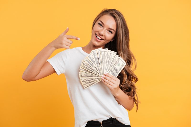 mulher sorrindo com um leque de dinheiro em uma mão e apontando para as notas com o dedo indicar da outra mão 