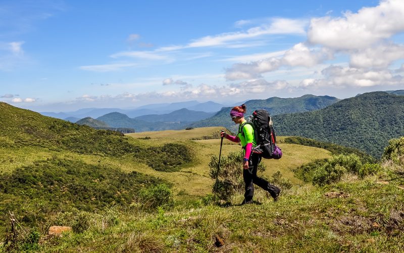 Foto de uma mulher com uma grande mochila nas costas e um cajado de caminhada nas mãos. Ela está caminhando no alto de um morro. Ao redor, vários outros morros verde. 