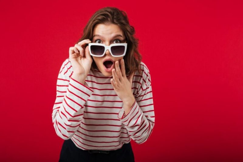 mulher de blusa branca listrada com vermelho, calça escura e o cabeça enrolado na altura do ombro; ela está com um óculos branco, segurando o mesmo com a mão direita e fazendo sinal de espanto com a boca e a mão esquerda 