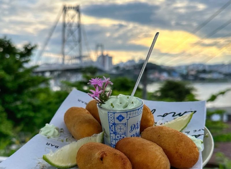 Foto de um prato com croquetes, limão fatiado e um copinho de maionese no meio. Ao fundo e de forma desfocada aparece a Ponte Hercílio Luz, o céu e algumas árvores. 