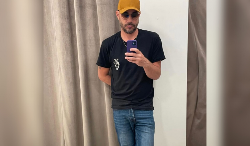 Evaristo Costa, apresentador - homem branco faz foto no espelho usando camiseta preta, calça jeans azul, boné cor mostarda e capa do celular roxa