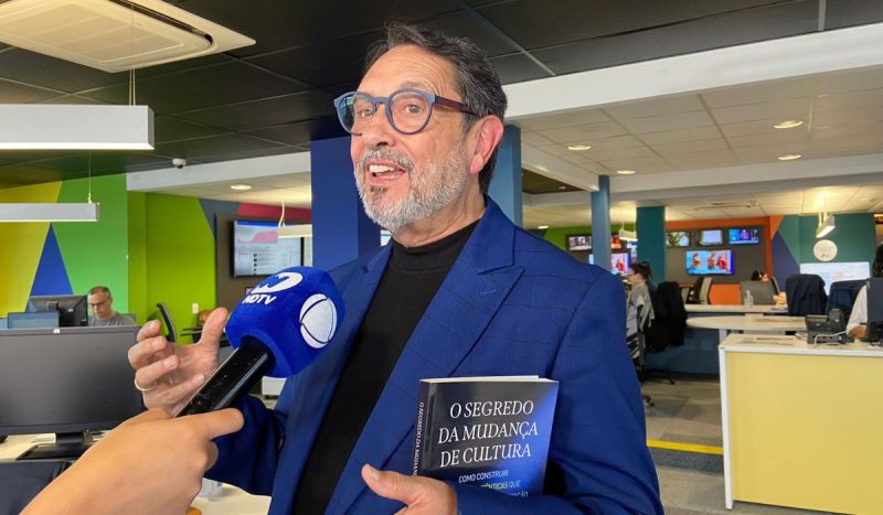 Autor de livro, homem branco, idoso e com barba e bigode grisalhos posa usando terno azul, segurando livro de capa azul e preto - ele fala sobre crescimento de empresas