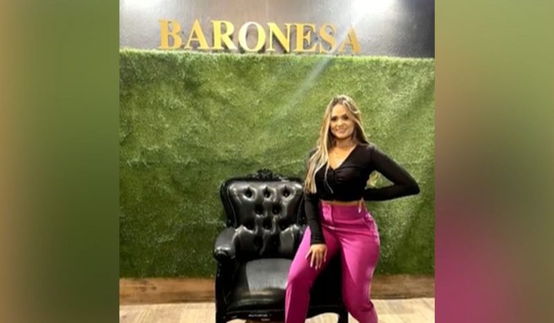 Francisca Alves da Silva, a baronesa do PCC - Mulher branca e loira, de cabelos compridos e lisos, sentada no braço de uma cadeira preta usando um top cropped de mangas na cor preta e uma calça na cor rosa. Ela está em frente a uma parede de grama verde com um letreiro acima dele com a palavra BARONESA escrita em letras maiúsculas