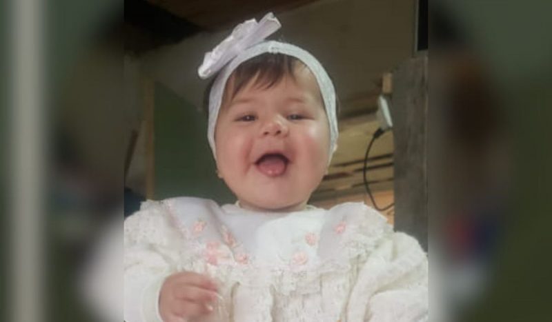 Bebê dada como morta em SC - menina, de 9 meses, com a boca aberta sorrindo sem dentes, ela tem os cabelos curtos e lisos e usa uma roupa branca com uma faixa de flor branca na cabeça