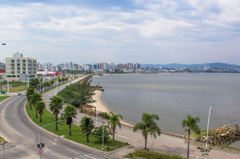 Do lado esquerdo da foto, aparece uma avenida, um canteiro central com várias árvores e uma pista de caminhada com um deck de madeira. No lado direito, as águas da Baía de São José. Ao fundo, vários prédios e morros verdes. O céu está azul. 