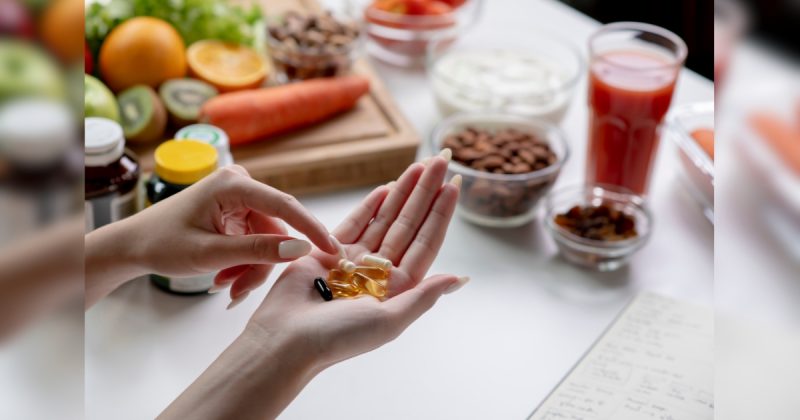 Mão segurando suplemento com copos e frutas por trás